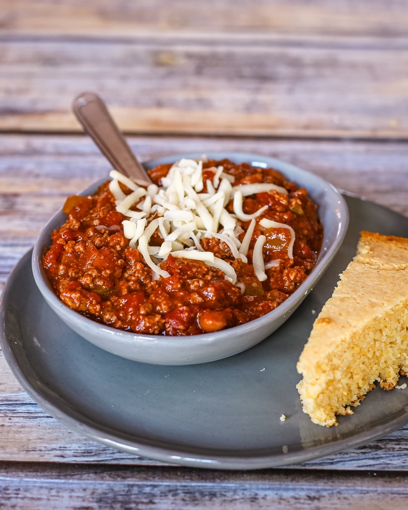 A serving of chili with some cheese and a side of cornbread.