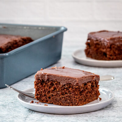 A slice of chocolate zucchini cake on a plate with the baking pan in the background.