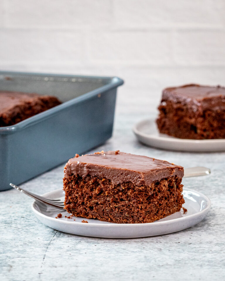 A slice of chocolate zucchini cake on a plate with the baking pan in the background.