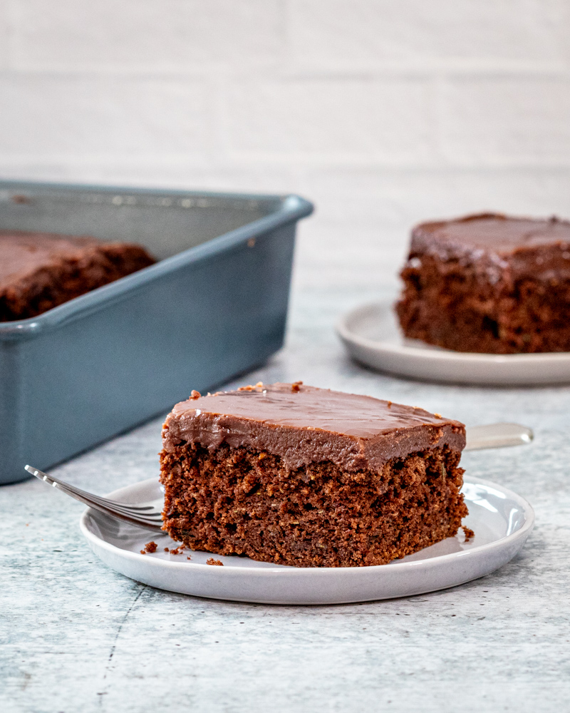 A slice of chocolate zucchini cake on a plate with the baking pan in the background.