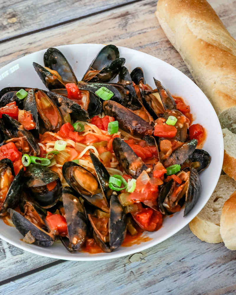 A bowl of mussels with Creole sauce, linguine, and a baguette on the side.