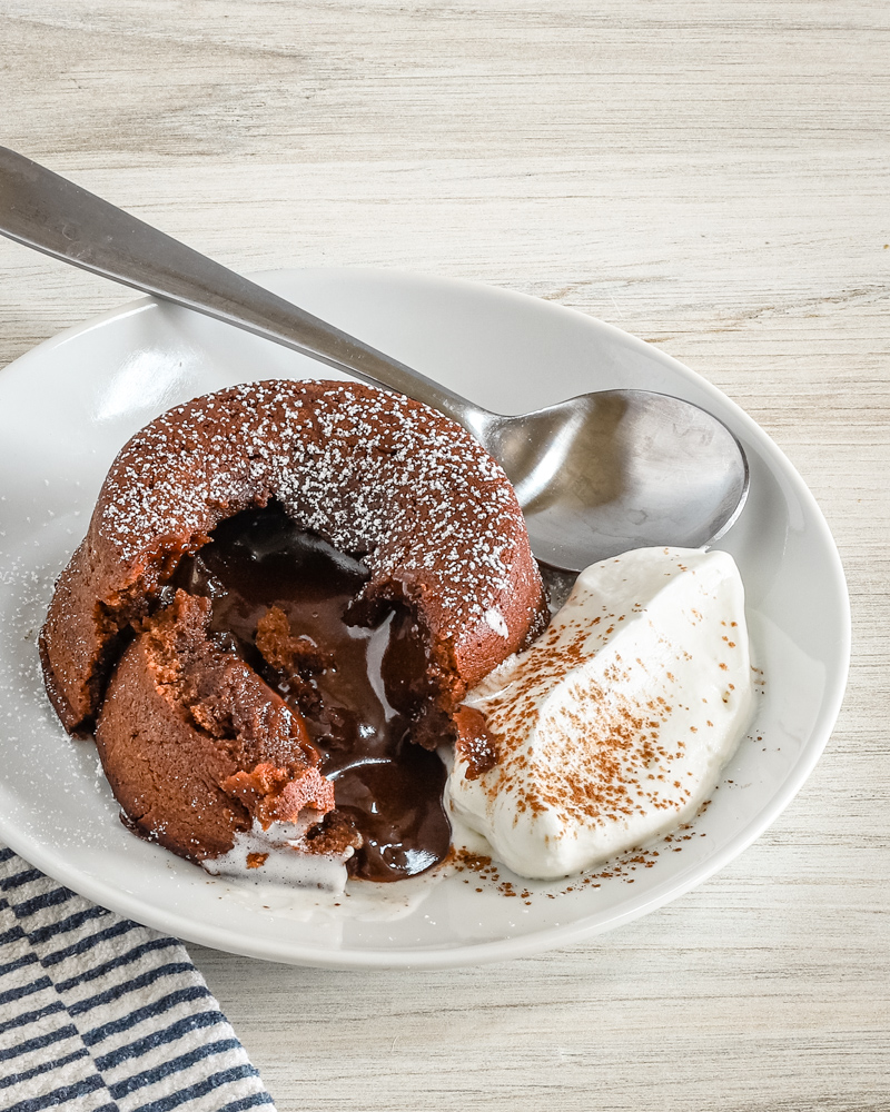 A chocolate fondant, also known as molten lava cake, on a plate with freshly whipped cream and a light dusting of cocoa and powdered sugar.