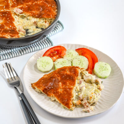 Ham and leek pie on a white plate with cucumbers and tomatoes, and the pie in the background.