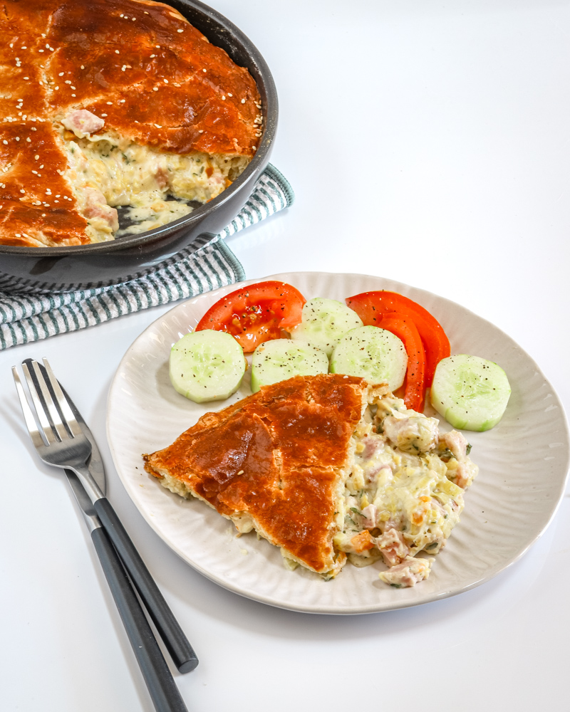 Ham and leek pie on a white plate with cucumbers and tomatoes, and the pie in the background.