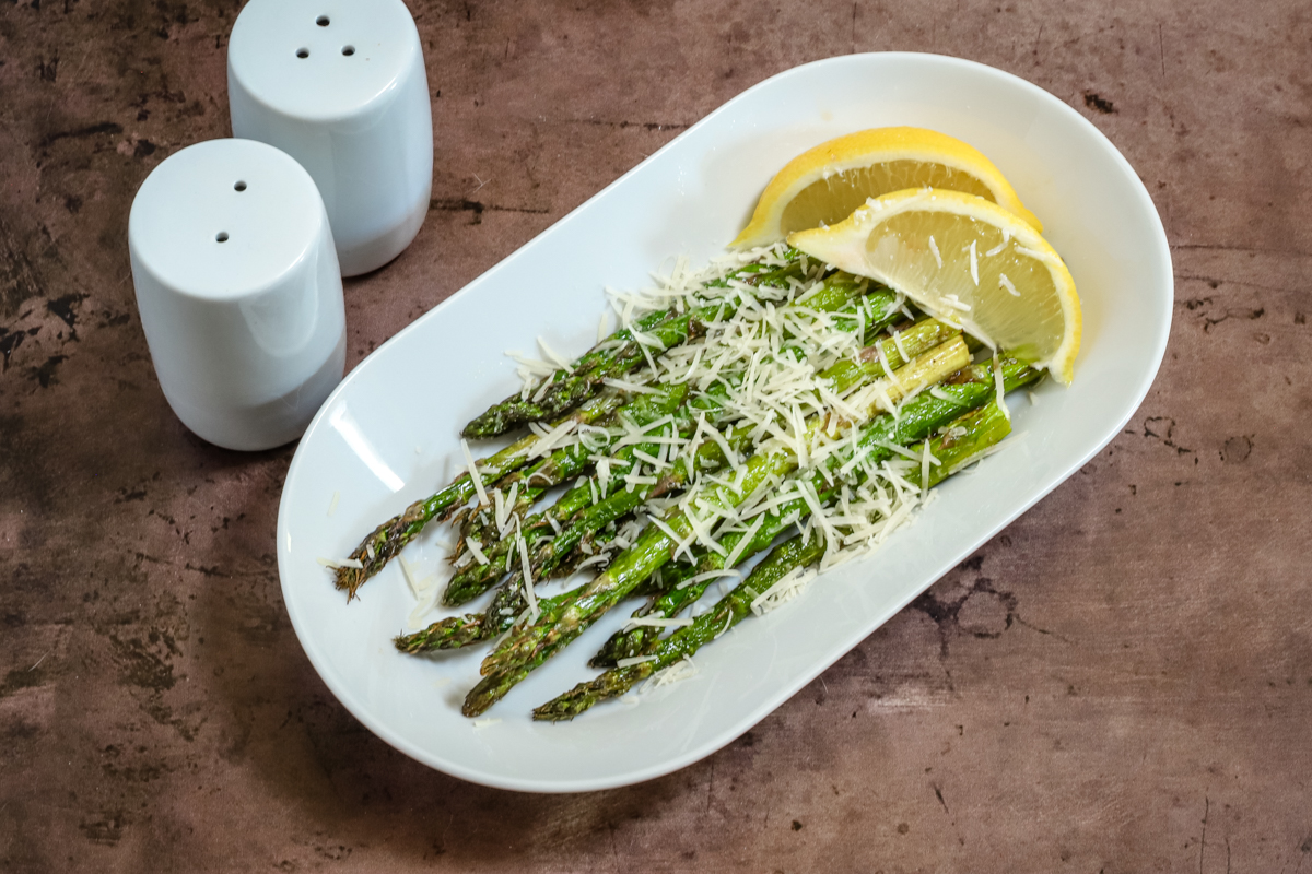 Air-fried asparagus in a serving dish with freshly grated Parmesan cheese and lemon wedges.