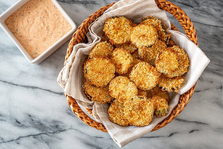Air fryer pickle slices in a basket with Sriracha mayo on the side.