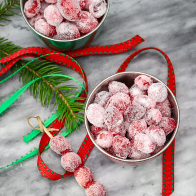 Sugared cranberries in small seasonal bowls and on skewers with festive holiday decorations scattered around them.