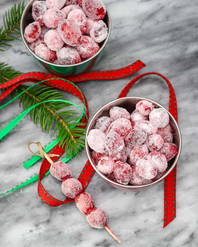 Sugared cranberries in small seasonal bowls and on skewers with festive holiday decorations scattered around them.
