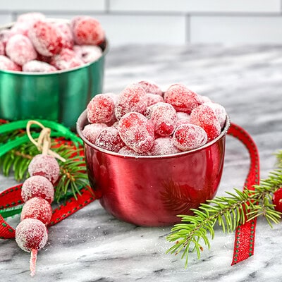 Sugared cranberries in small seasonal bowls and on skewers with festive holiday decorations scattered around them.