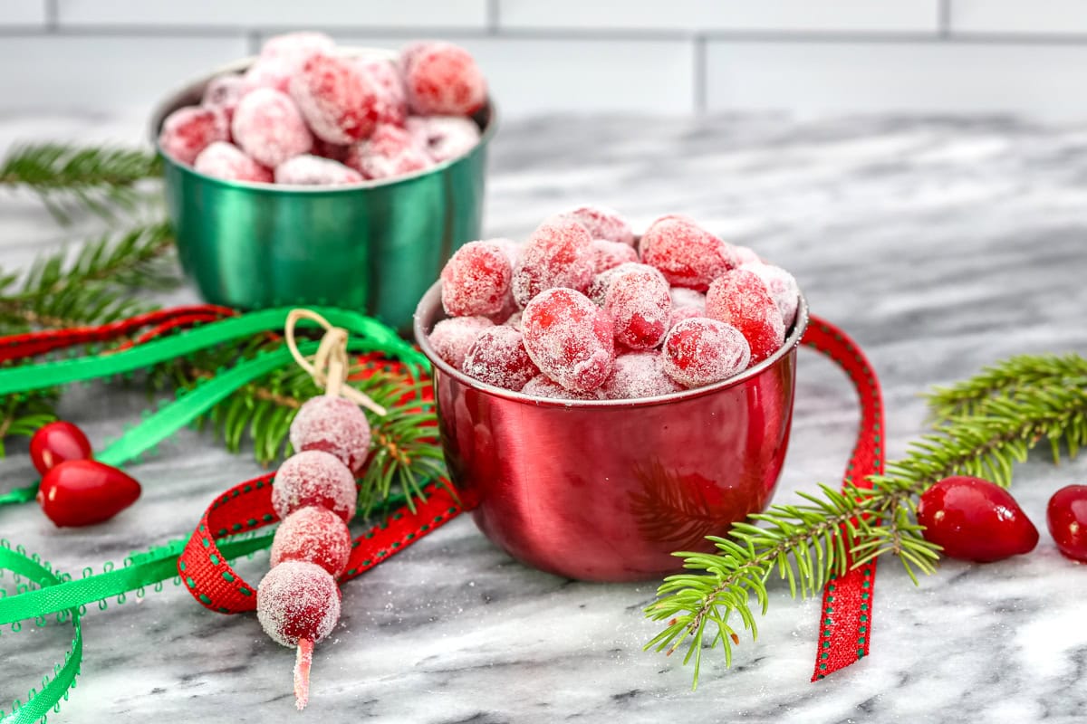 Sugared cranberries in small seasonal bowls and on skewers with festive holiday decorations scattered around them.