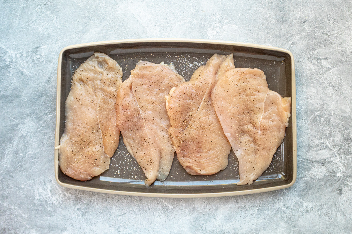 Thin chicken cutlets on a tray, seasoned with salt and pepper.
