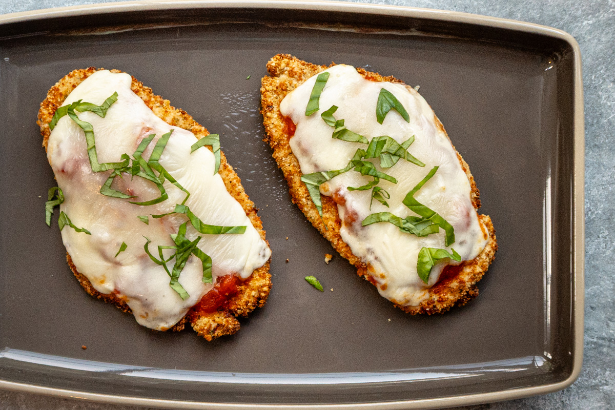 A closeup look at two air fried chicken breast cutlets topped with tomato marinara sauce, mozzarella cheese, and fresh basil.