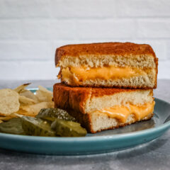 A grilled cheese sandwich from the air fryer, sliced and arranged on a plate with pickles and potato chips.