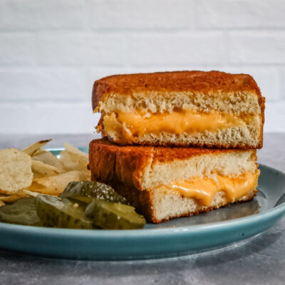 A grilled cheese sandwich from the air fryer, sliced and arranged on a plate with pickles and potato chips.