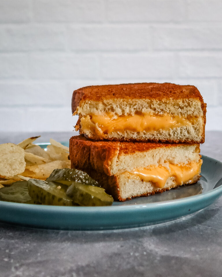 A grilled cheese sandwich from the air fryer, sliced and arranged on a plate with pickles and potato chips.