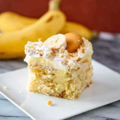 A banana pudding poke cake with a garnish of sliced banana and vanilla wafer, and bananas in the background.