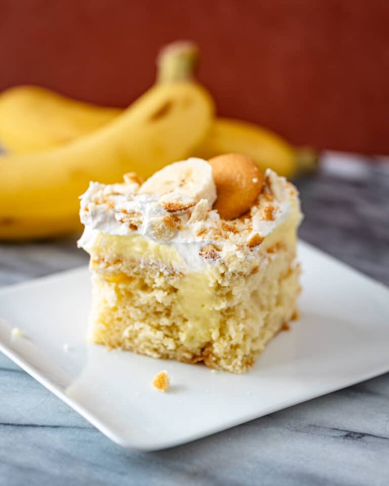 A banana pudding poke cake with a garnish of sliced banana and vanilla wafer, and bananas in the background.
