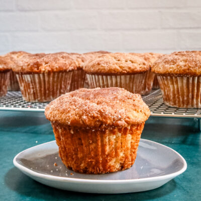 A cinnamon muffin on a small plate with the rack of muffins behind it.