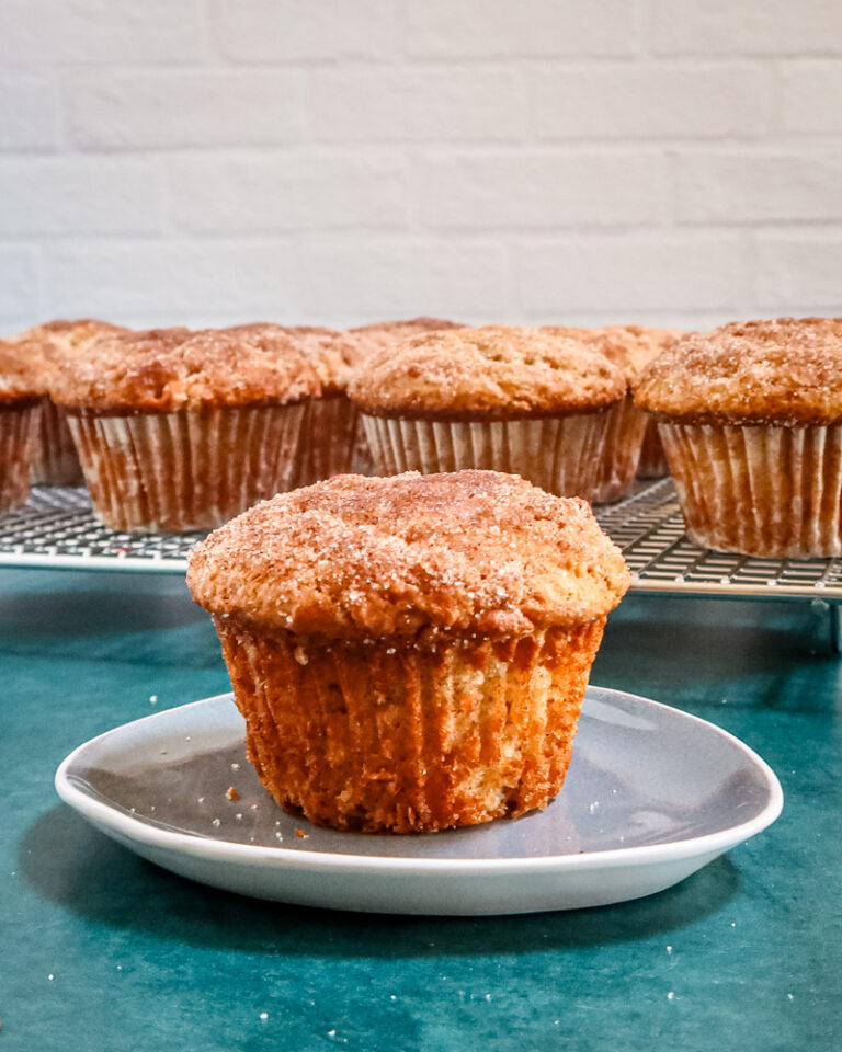 A cinnamon muffin on a small plate with the rack of muffins behind it.