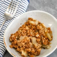A serving of apple crisp in a dessert dish with a striped cloth and fork beside it.
