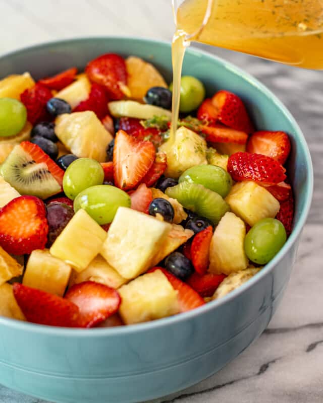 A honey citrus dressing being poured onto a fresh fruit salad in a serving bowl.