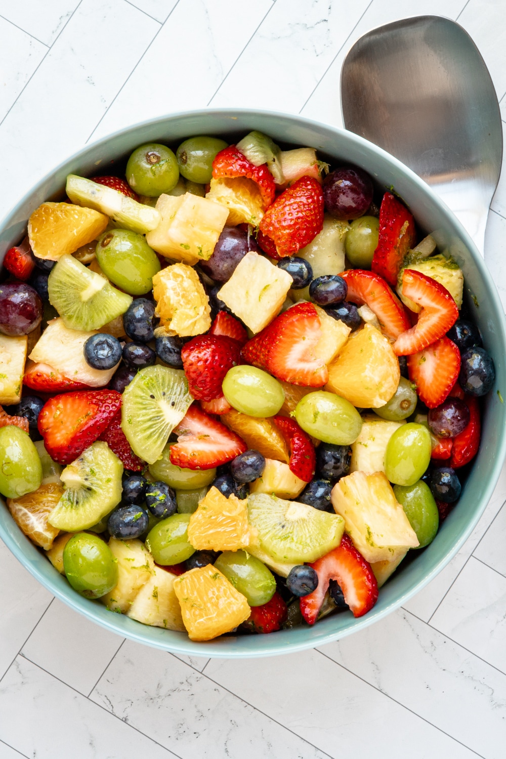 A fresh fruit salad in a large serving bowl with a large serving spoon to the side.