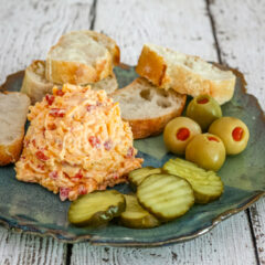 Pimiento cheese on a plate with olives, pickles, and baguette slices.