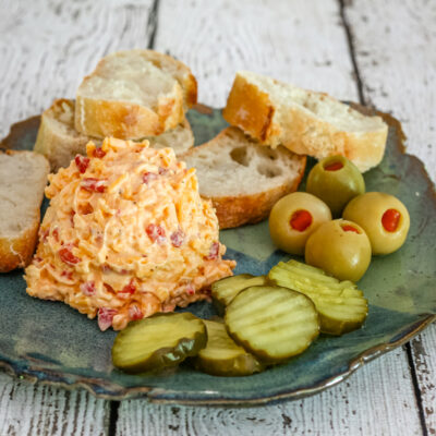 Pimiento cheese on a plate with olives, pickles, and baguette slices.