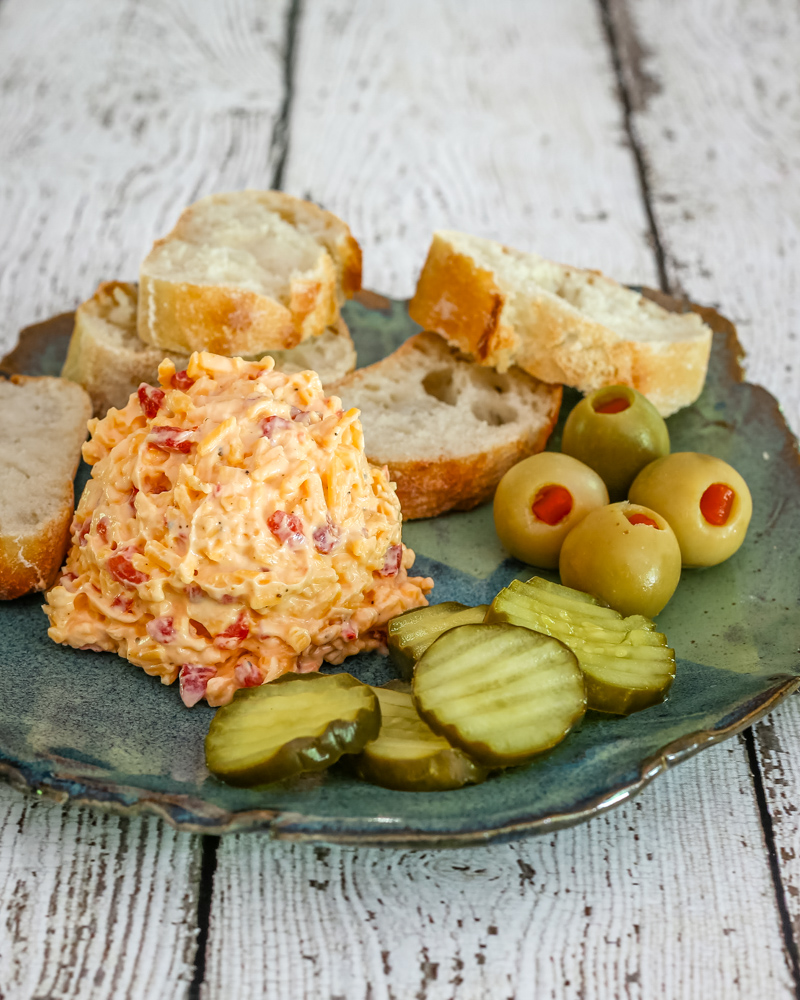 Pimiento cheese on a plate with olives, pickles, and baguette slices.