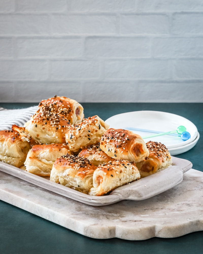A serving tray with baked puff pastry pigs in a blanket with small appetizer plates and picks for serving.