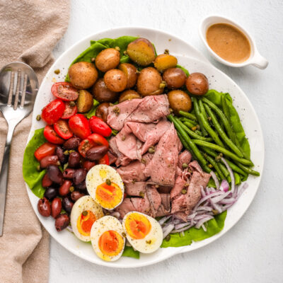 Roast beef nicoise salad arranged on a platter with eggs, deli sliced roast beef, tomatoes, olives, potatoes, onions, green beans, and a Dijon mustard dressing.