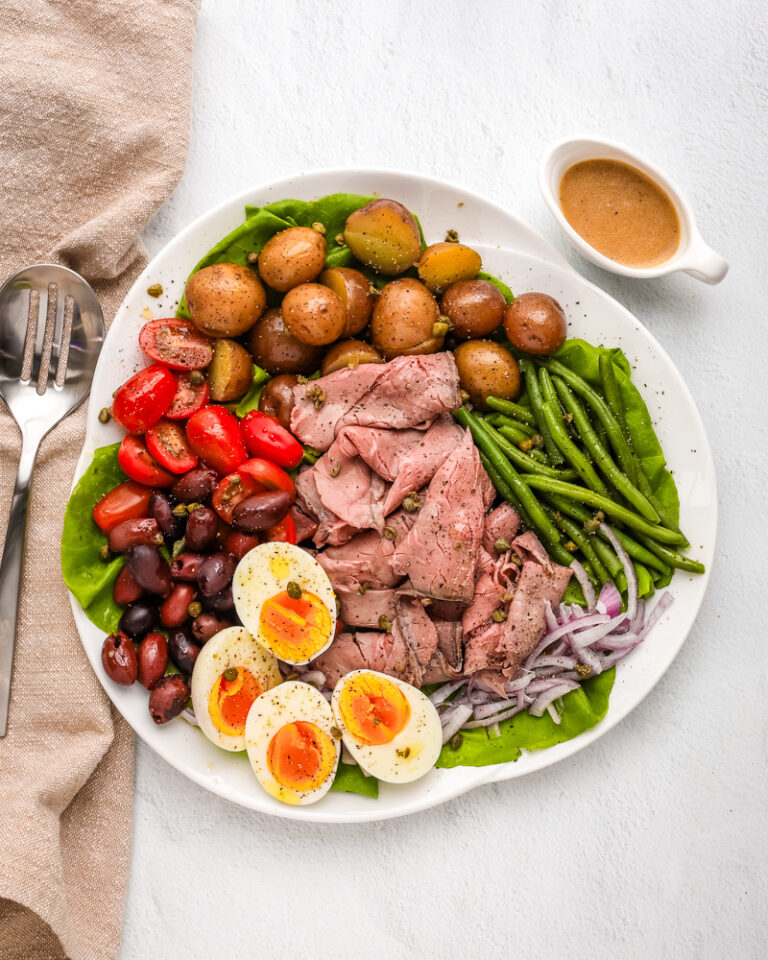 Roast beef nicoise salad arranged on a platter with eggs, deli sliced roast beef, tomatoes, olives, potatoes, onions, green beans, and a Dijon mustard dressing.