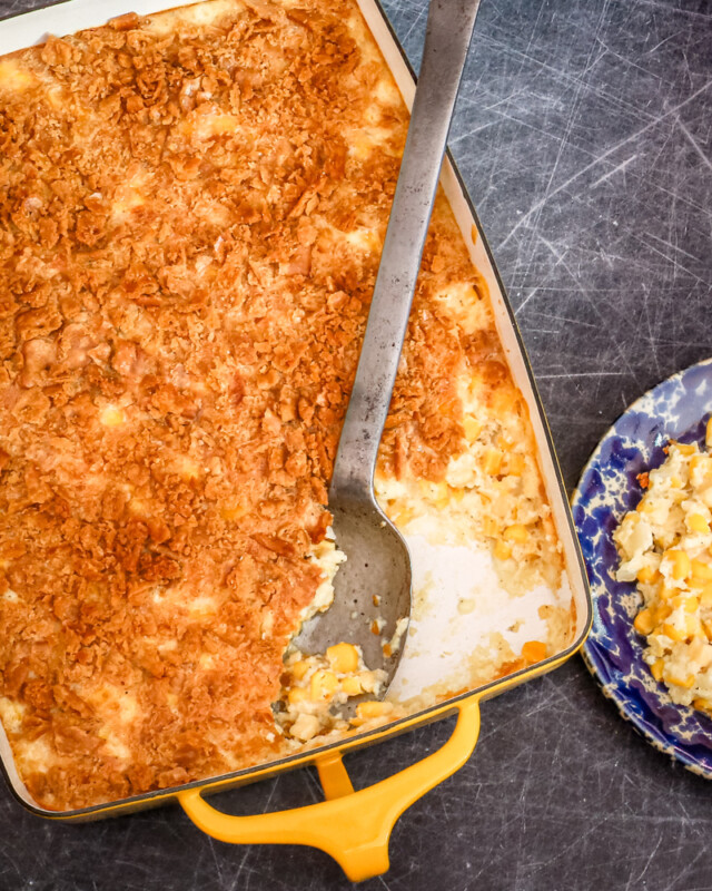 A scalloped corn casserole in a yellow baking dish with a large serving spoon.
