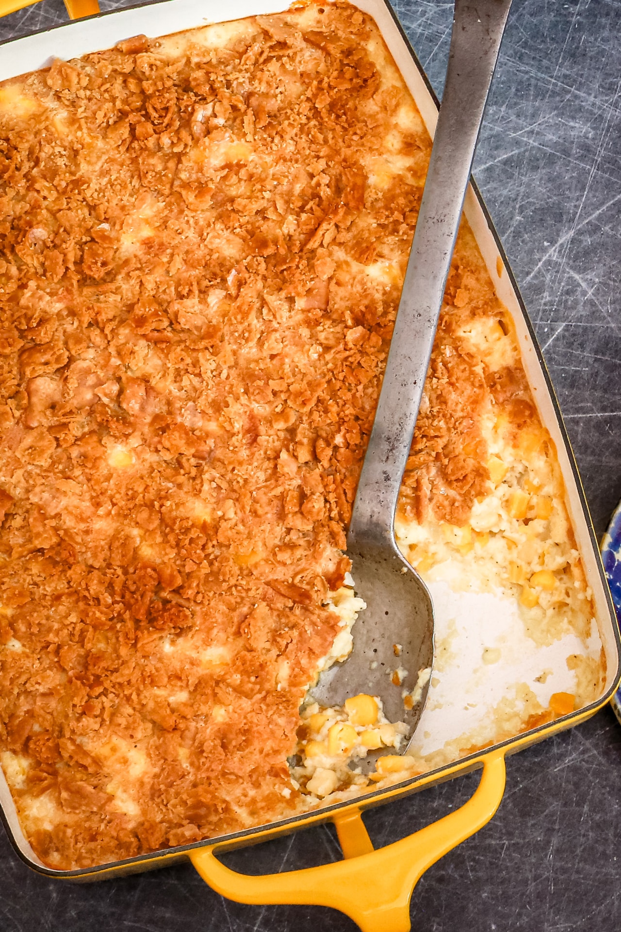 A scalloped corn casserole in a yellow baking dish with a large serving spoon.