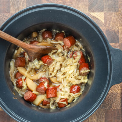 Slow cooker cooked kielbasa with apples, cabbage, onions, and tangy sauce, shown in the crock pot with a wooden spoon.