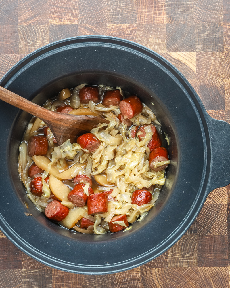 Slow cooker cooked kielbasa with apples, cabbage, onions, and tangy sauce, shown in the crock pot with a wooden spoon.