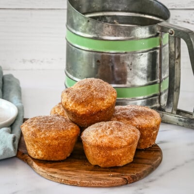 Snickerdoodle muffins on a small cutting board with a flour sifter in the background.