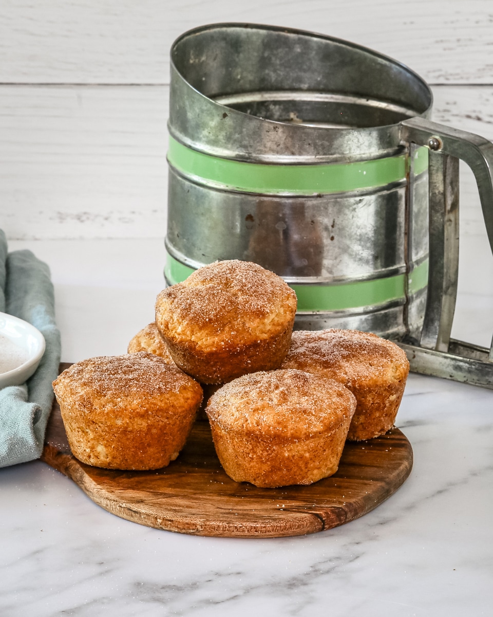 Snickerdoodle muffins on a small cutting board with a flour sifter in the background.