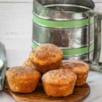Snickerdoodle muffins stacked on a small cutting board with a flour sifter behind it and some cinnamon sugar to the side.