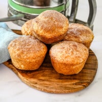 Snickerdoodle muffins stacked on a small cutting board with a flour sifter behind it and some cinnamon sugar to the side.