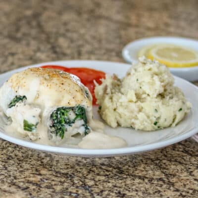A rolled-up stuffed flounder fillet with shrimp stuffing on a white plate with rustic mashed potatoes, tomatoes, and lemons.