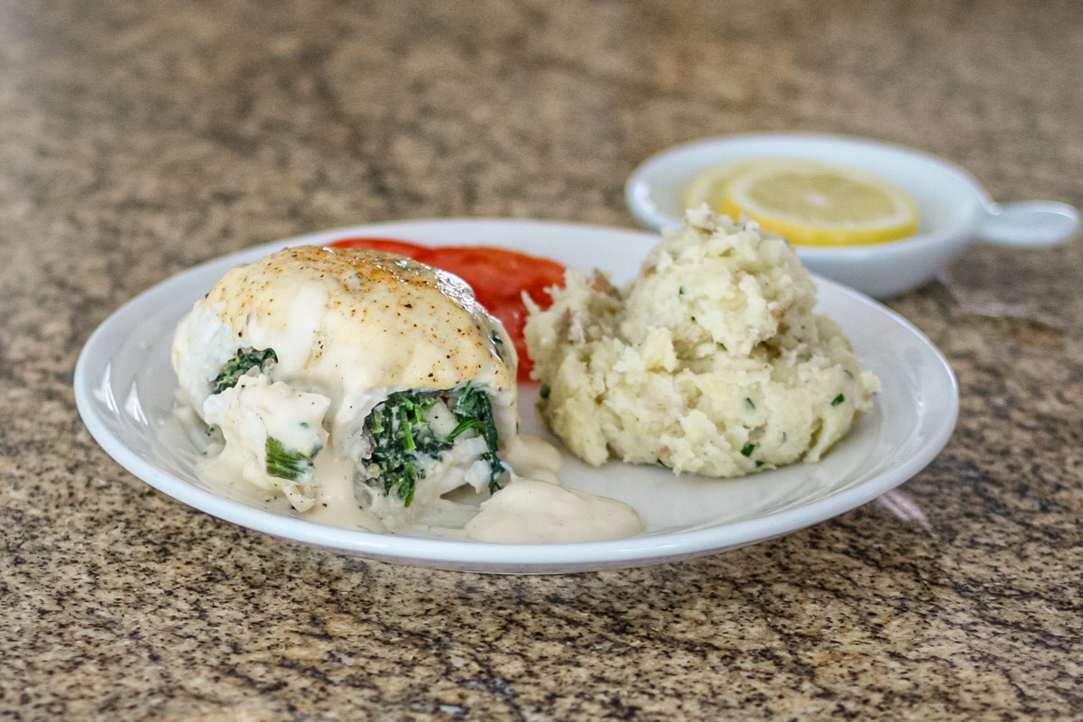 A rolled-up stuffed flounder fillet with shrimp stuffing on a white plate with rustic mashed potatoes, tomatoes, and lemons.