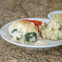 A rolled-up stuffed flounder fillet with shrimp stuffing on a white plate with rustic mashed potatoes, tomatoes, and lemons.