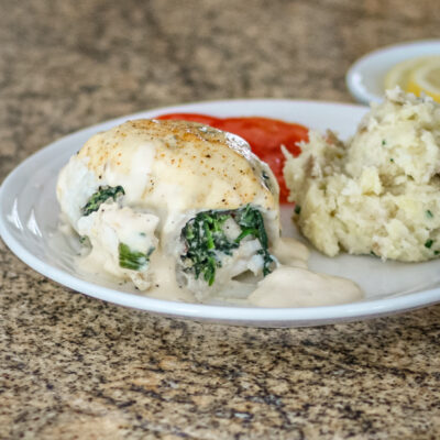 Stuffed flounder on a plate with mashed potatoes, tomato slices, and lemon in the background.
