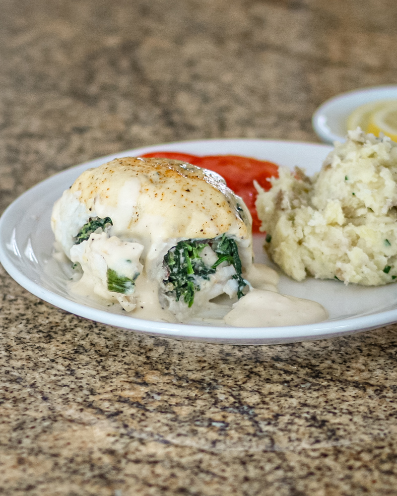 Stuffed flounder on a plate with mashed potatoes, tomato slices, and lemon in the background.