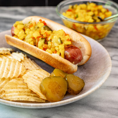 Corn relish on a hot dog with some chips and pickles on the side.