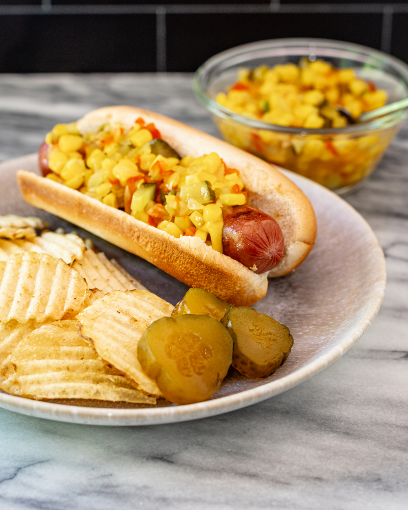 Corn relish on a hot dog with some chips and pickles on the side.