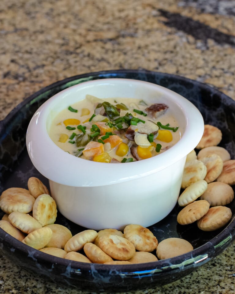 Creamy vegetable and sausage chowder shown in a bowl with chives for garnish and oyster crackers.