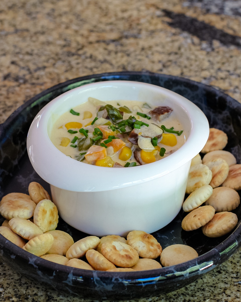 Creamy vegetable and sausage chowder shown in a bowl with chives for garnish and oyster crackers.