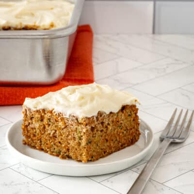 A zucchini carrot cake slice on a white plate with cream cheese frosting.